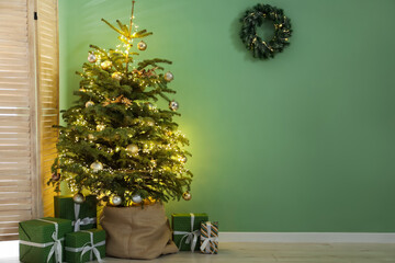 Fototapeta premium Beautiful fir tree with Christmas decor in pot, gift boxes and folding screen near green wall indoors. Space for text