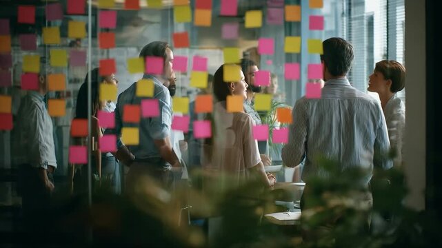 Team brainstorming in modern office, seen through a glass wall covered with colorful sticky notes. Collaboration, business planning, creativity, teamwork, innovation, diverse workplace, and strategy