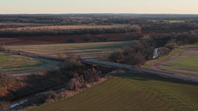 Aerial drone pull back and tilt up of a car driving across a rural bridge over a creek through farmland in early spring at sunset in Oklahoma.