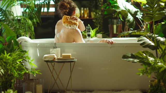 A woman with a bun takes a bath, using a loofah on her back. Surrounded by green plants, the bathroom offers a botanical, greenhouse-like atmosphere, ideal for tranquil relaxation & self-care.