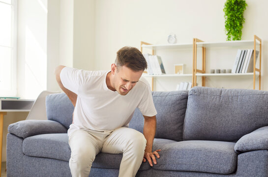 Sick man with lower back pain sitting on sofa at home. Unhappy young person suffering from backache and lumbago, chronic spine orthopedic disease of herniated disc or muscle spasm while moving