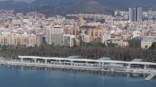 Aerial view of Puerto de Malaga harbor with Muelle Uno pergola, La Manquita with scaffolding, palm trees, parkland, twin towers, mountains, boats and service vehicles.