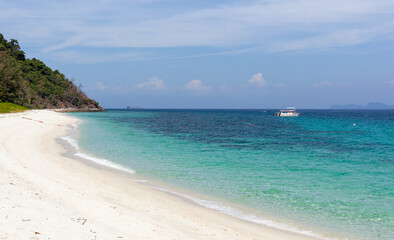 Fototapeta premium Tropical Koh Adang Beach in Thailand. White sand shoreline with turquoise water and clear blue sky. Boat in distance with lush green hills nearby.