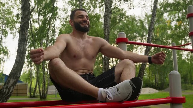 A happy, shirtless, bearded man with a muscular build sits in a lotus pose on red parallel bars at an outdoor gym, meditating and smiling after a workout. He is surrounded by green trees.