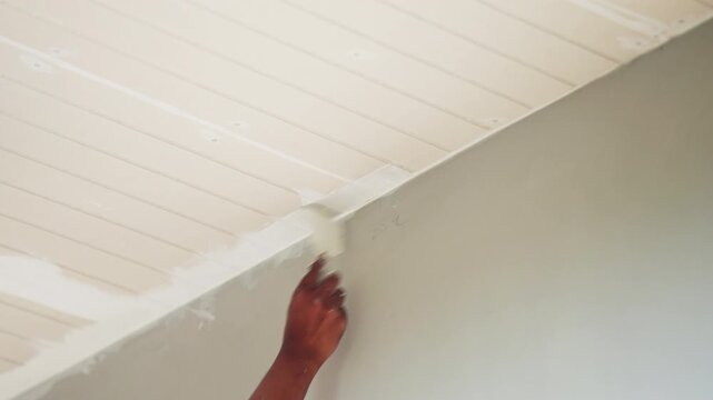 Close up of worker hand painting white ceiling with brush during home renovation. Interior improvement, house maintenance, DIY painting and construction work concept in residential building.