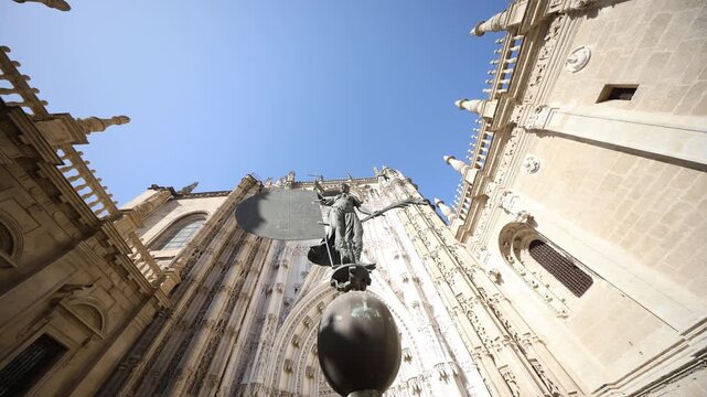 Low angle view in Seville, Andalusia shows Gothic facade, Giraldillo vane on orb, carved saints and tracery. Midday light casts crisp shadows as camera tilts upward.