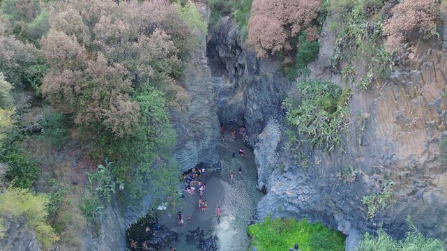 Cinematic aerial drone panorama of Alcantara Gorges in Sicily showing the dramatic lava canyon carved by the Alcantara River with basalt rock formations and crystal clear water flowing through a uniqu