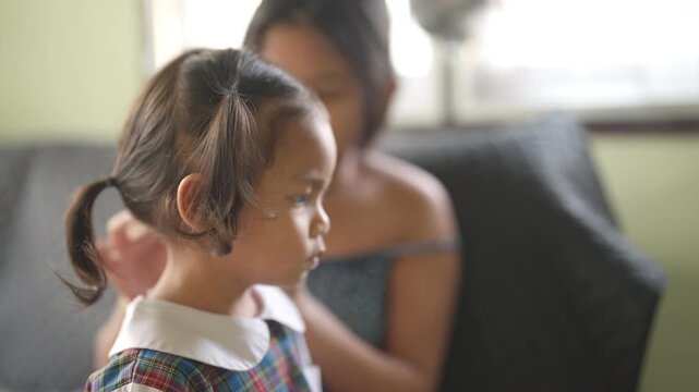 Asian mother braiding young daughter hair inside living room. Family lifestyle, parenting care and loving relationship moment between mother and child.