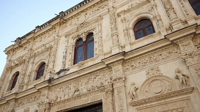 Close detail of a Spanish Plateresque civic facade as the camera pans upward diagonally, showing sculptural bands, cherubic figures, medallions, and stained glass at midday.