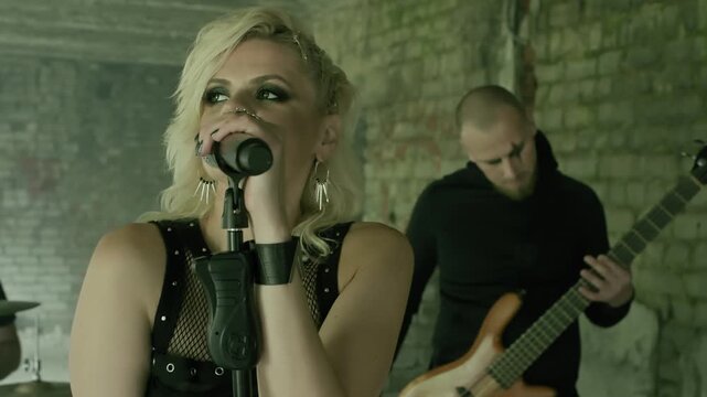 Female rock singer with blonde hair and dramatic makeup sings passionately into a microphone. A bassist plays in the background in a gritty, abandoned building. Rock band music video.