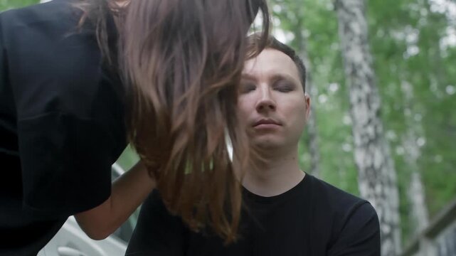 A makeup artist applies dark eyeshadow to a man's closed eye. The man sits patiently outdoors in a forest, preparing for a photoshoot or performance with a goth or rock style aesthetic.