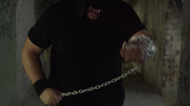 A mysterious man in a black hoodie stands in a derelict corridor, aggressively wrapping a heavy metal chain around his arm in preparation for a fight. Dark, gritty, and intimidating atmosphere.