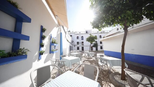 Cobalt blue frames, wall planters, cobblestones, gingham tables, and orange trees sit empty as a slow pan shows lens flare, sharp shadows, and subtle leaf movement.
