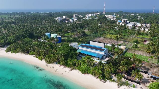 Aerial drone view of a water desalination plant with large blue storage tanks on the coast of Thoddoo Island, Maldives, providing fresh water via reverse osmosis.