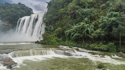 A picturesque tropical waterfall. Streams of white water flow down from the cliff. Foam, splashes, fog. Rapids in the riverbed. Lush green vegetation. China. Huangguoshu Waterfall. Guizhou