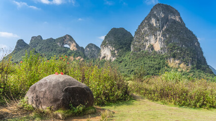 Picturesque karst mountains, the blue sky. There is a rounded arched opening at the top. Green vegetation on steep slopes. In the foreground is a boulder on the lawn. Red hibiscus flowers on a stone   © Вера 
