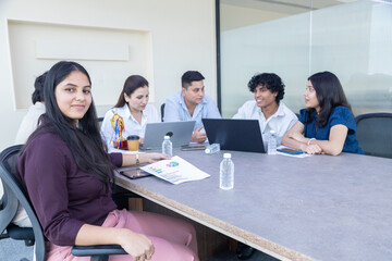 Fototapeta premium Happy Indian business woman employee sitting with other office colleagues in meeting room working together on laptop, sales and company growth report, Corporate job. Planning, strategy and Teamwork