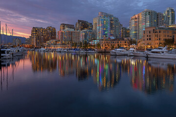Obraz premium Luxury city skyline marina with modern buildings and boats reflecting colorful lights on calm water at sunset, creating peaceful and vibrant urban atmosphere