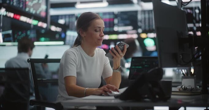 While Monitoring Real Time Stock Indices, Tech Savvy Female Specialist Records Voice Command or Update On Mobile Phone. Efficiency of Instant Audio Communication in Sophisticated Exchange Hub.