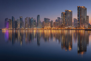 Fototapeta premium Modern waterfront city skyline at blue hour with illuminated skyscraper reflections on calm water creating peaceful and vibrant urban atmosphere