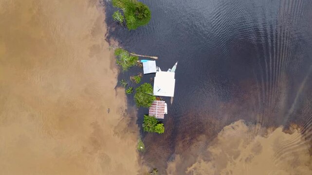 This aerial shot shows a small house surrounded by dark water and light brown riverbanks in Leticia, Colombia.