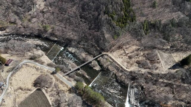 Rugged river flows through forested valley. Small dam and bridge span the rushing waters. Furrows mark fields beside the stream. Quiet, natural beauty of remote countryside. Perfect for environmental
