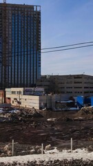 Fototapeta premium Urban construction site with exposed earth and partial snow in city. Tall skyscraper building under urban construction, rising above older city structures and power lines.