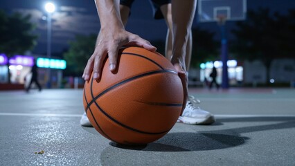 Naklejka premium Close-up of a basketball player's hands gripping an orange basketball on a court at night, with dynamic lighting and a blurred background showing spectators.