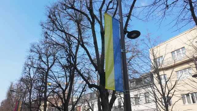A vibrant urban street scene with a Ukrainian flag waving under clear blue skies. The bare trees and classical architecture add charm and suggest a chilly winter morning, captured in this static shot.
