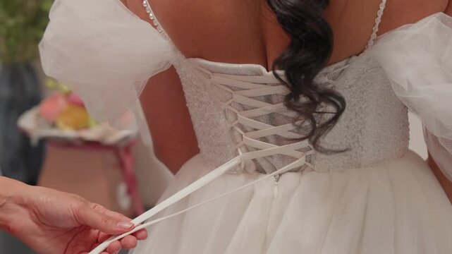 Detailed view of a bride's dress being laced up on her wedding day. The intricate lacework and delicate fabric showcase elegance and romance in soft, ambient lighting. Perfect for wedding themes.