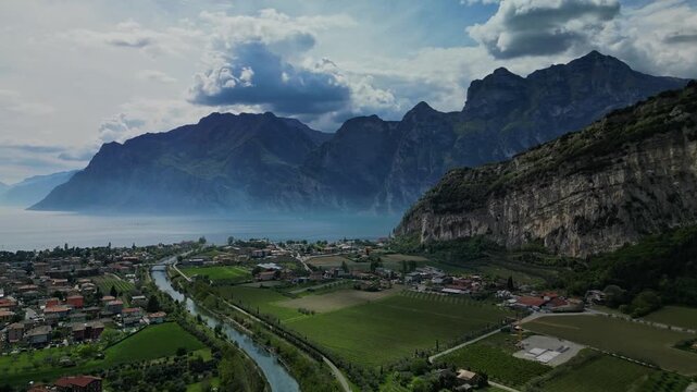 Aerial drone view flying over Sarca River valley toward Lake Garda near Nago-Torbole in northern Italy, village houses and dramatic alpine cliffs under a cloudy sky.
