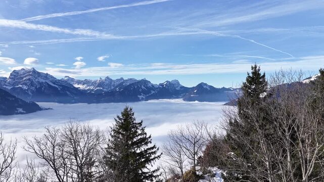 Aerial revealing view of Walensee in Switzerland as the lake is covered by fog, and the surrounding alpine mountains emerge above a dramatic sea of clouds beneath a bright blue sky.