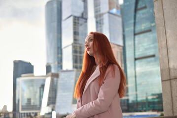 Fototapeta premium A red-haired woman stands in front of modern glass skyscrapers. She is wearing a light pink coat and is looking directly at the camera with a calm, composed expression.