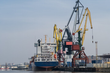 Fototapeta premium Cargo ship docked at Kaliningrad port with colorful cranes and shipping containers visible along the waterfront under a clear sky