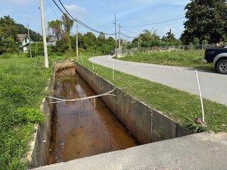 A roadside drainage ditch with a shallow stream of water flowing through it. The low water level suggests dry weather conditions or reduced drainage flow within a simple roadside setting.