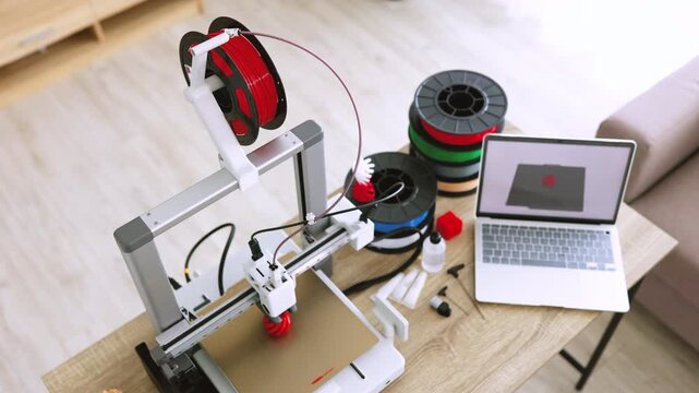 A 3D printer is actively creating an object with red filament, alongside spools of filament and a laptop displaying the design