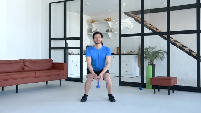 Motivated handsome active fit man in sportswear practicing weight training, doing dumbbell sumo squats exercise improving lower body strength while working out in domestic room.