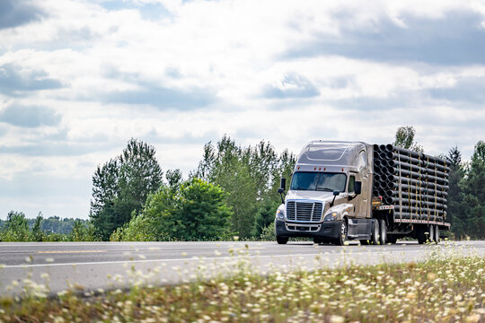 Beige pro big rig semi truck transporting fastened plastic pipes on the step deck semi trailer driving on the summer highway road