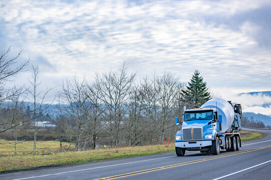 Blue big rig day cab semi truck with mixer trailer application transporting mixed concrete running on the highway road