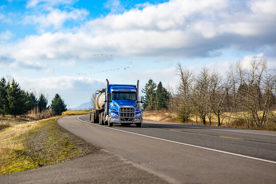 Bright blue day cab big rig semi truck tractor transporting liquid cargo in tank semi trailer driving on the flat highway road at sunny day