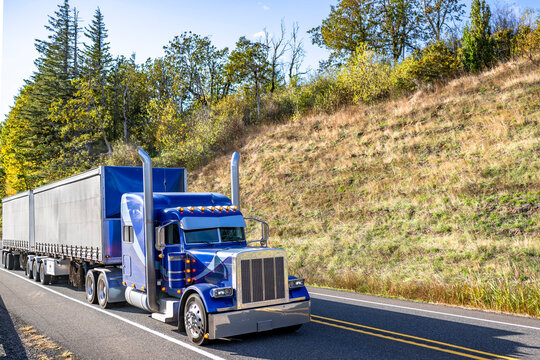 Bright ultramarine blue classic big rig semi truck tractor with chrome parts transporting cargo in conestoga frame flat bed semi trailers running on the mountain road with autumn trees on the hill