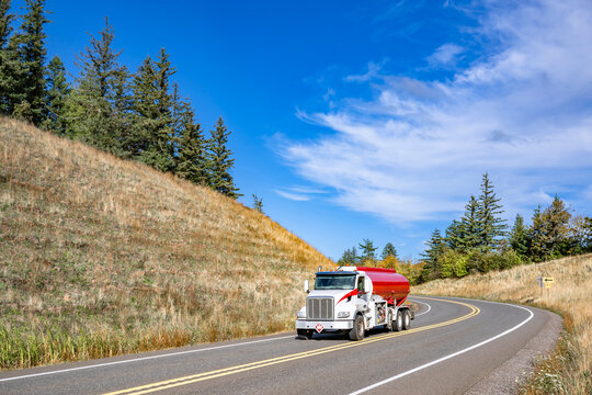 Day cab white big rig semi truck with red tank semi trailer transporting fuel running on the winding mountain road with dry grass on the hills
