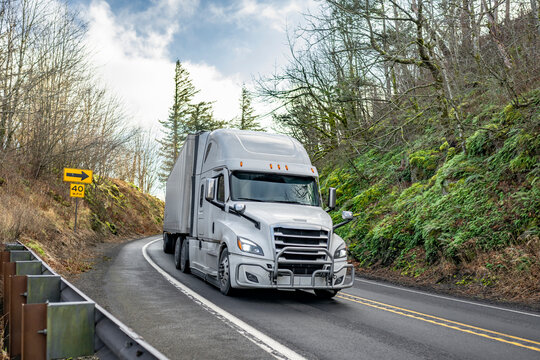 Industrial white bonnet big rig semi truck with grille guard transporting cargo in dry van semi trailer running on the winding narrow road with forest on hills