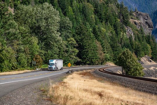 Day cab big rig semi truck with green roof spoiler transporting cargo in bulk semi trailer running on the winding road along the rail road in Columbia Gorge area