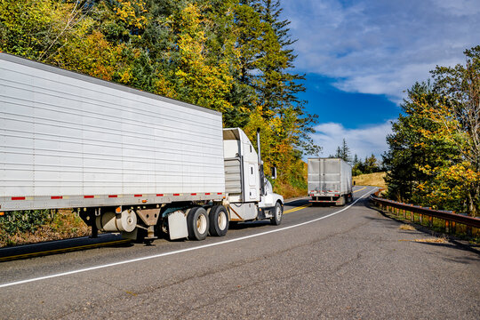 Powerful white big rig semi truck tractor with extended cab transporting cargo in refrigerated semi trailer running on the autumn winding freeway road behind another truck