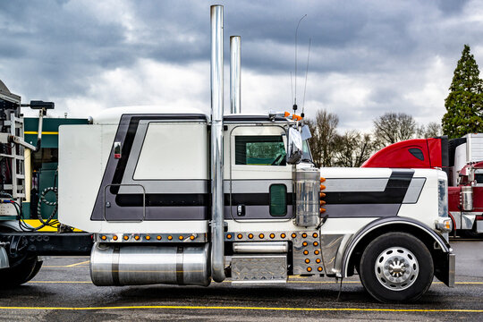 Profile of the classic American big rig white semi truck with high exhaust pipes and bulk semi trailer standing on the truck stop parking lot take a break