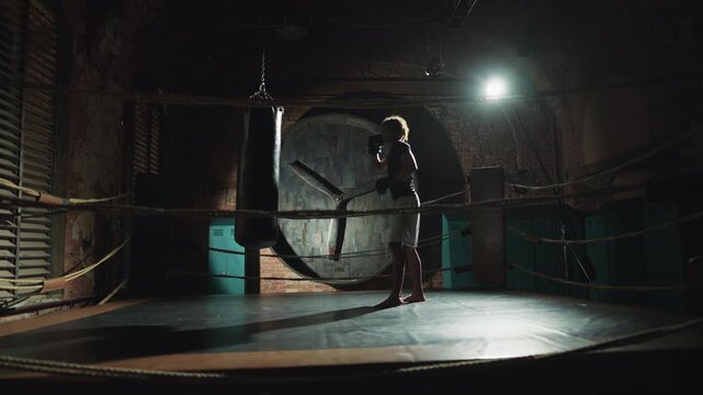 African american athlete practicing box in dark gym, hitting punching bag. Kickboxing and combat sports, self-defense class for men, professional sportsman training alone in night, future champion