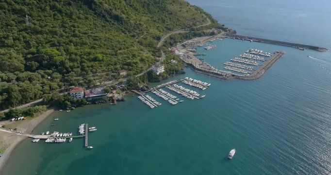 Aerial view of a bustling marina filled with white yachts tucked into a turquoise bay. Lush green slopes meet the deep blue sea, creating a stunning contrast. It's the Sapri port, in Campania, Italy.