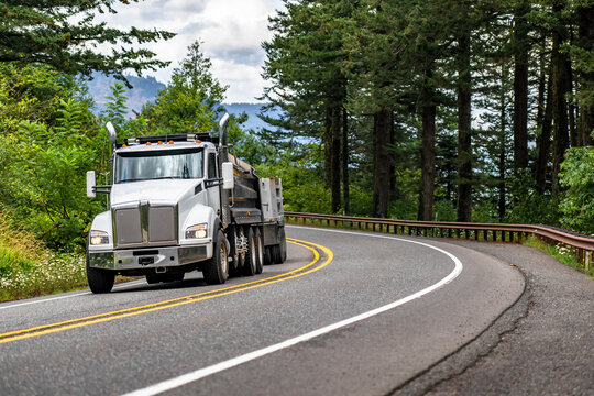 White tip big rig semi truck with two tipper trailers running on the winding mountain road with trees on the sides