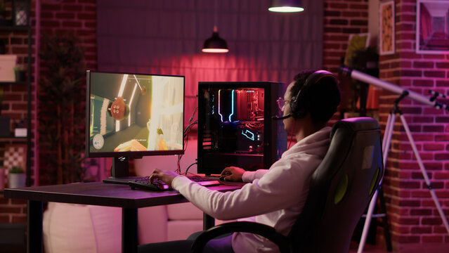 Static tripod shot of african american gamer streaming first person shooter while explaining gameplay while using pc gaming setup. Man relaxing playing online action game talking to team on headset.